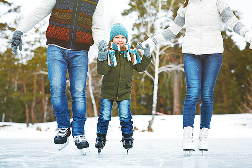 Portrait of happy little boy in winterwear , holding hands of his parents and having fun on winter ice rink surrounded by trees, blurred background