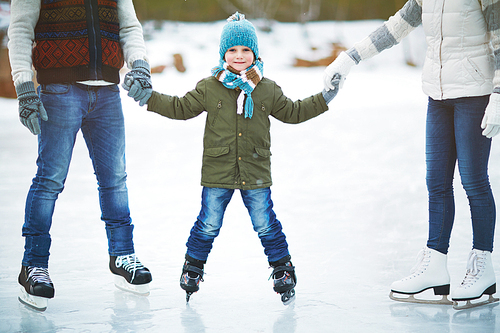 Portrait of rosy-cheeked little boy with smile on his face holding hands of his parents and enjoying winter day on outdoor skating rink, blurred background