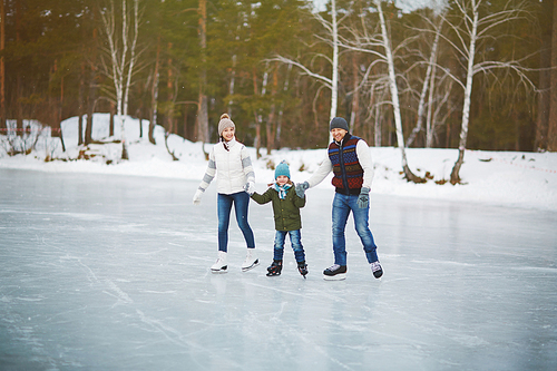 Portrait of cheerful family of three in winterwear , holding hands and spending their weekends on winter ice rink surrounded by forest