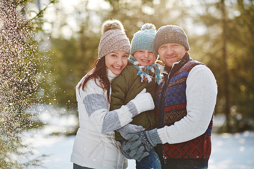 Portrait of lovely family of three with smiles on their faces and snowflakes on their hats and outwear posing in snowy winter park, blurred background