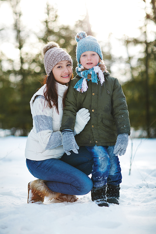 Portrait of rosy-cheeked little boy with snow on his outwear and his attractive young mother sitting on haunches and embracing him slightly in snowy winter park, blurred background