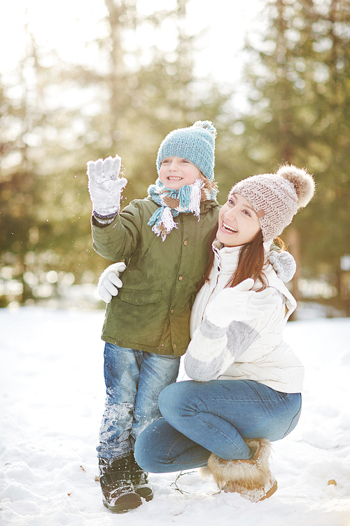 Portrait of happy family: attractive young mother sitting on haunches and hugging gently her little son trying to catch snowflakes in snowy park on sunny winter day