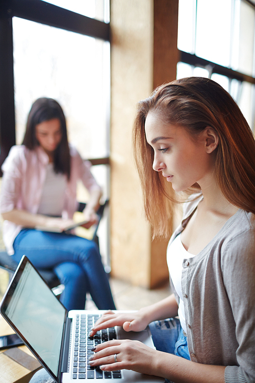 Youthful girl typing on laptop with her friend on background