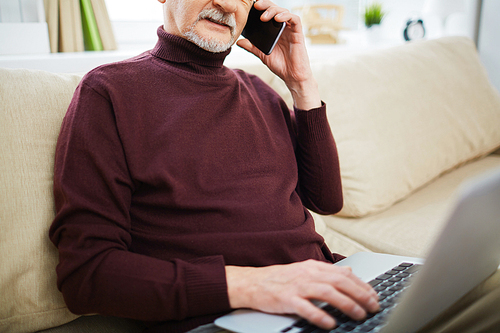 Senior man sitting on sofa and using his laptop and talking on the phone
