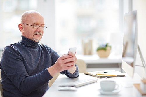 Mature man with cellphone sitting by computer in office