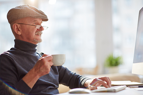 Senior man in cap drinking coffee and typing on keyboard