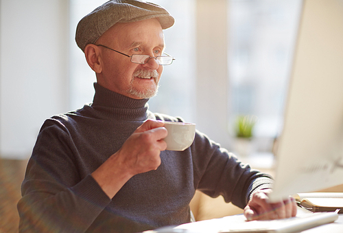 Happy mature man in cap, eyeglasses and pullover drinking tea in front of computer