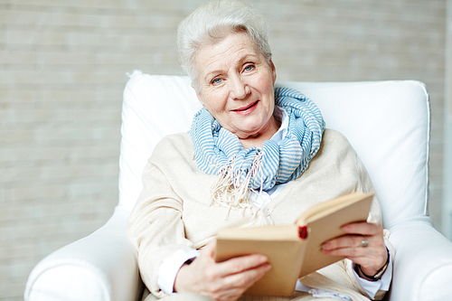 Happy senior female sitting in arm-chair with open book and 