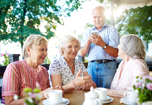 Happy aged women talking by table with senior man with cellphone standing near by
