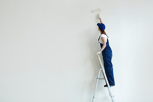 Female in coverall painting wall while standing on ladder