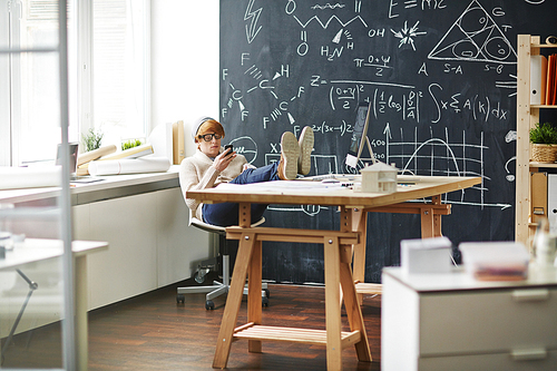 Young designer or architect with smartphone having break in office