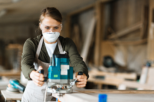 Young female in uniform, protective eyeglasses and respirator woodworking with electric fretsaw