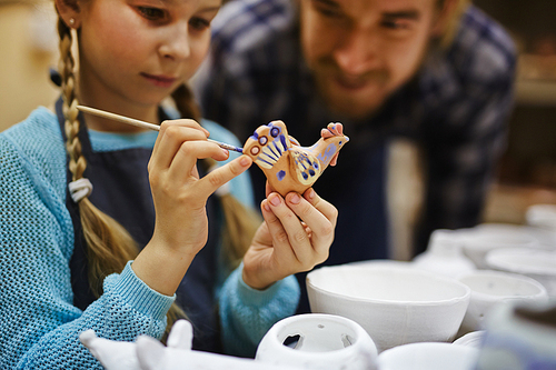 Little girl painting clay bird in workshop of her father