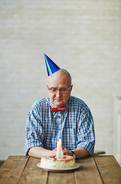 Mature man looking at birthday cake with burning candle on table