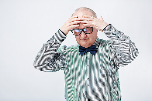 Waist up portrait of dressed up senior man in glasses and bowtie looking stressed and rubbing his head with hands isolated on white