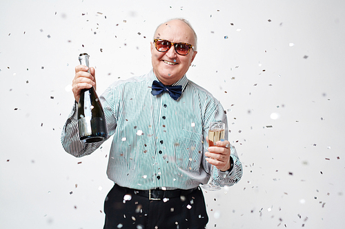 Waist up portrait of smiling well-dressed senior man in sunglasses holding bottle of sparkling wine and full wineglass with confetti falling from above against white background.