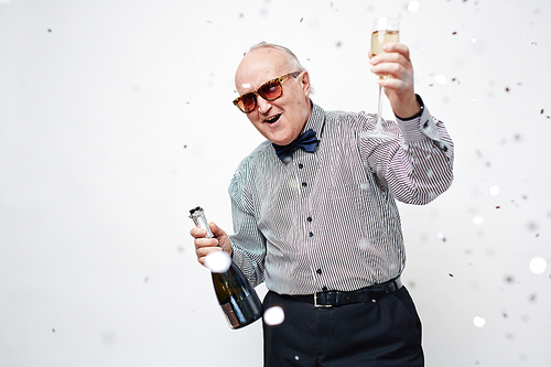 Waist up portrait of senior man in sunglasses dressed in stylish shirt with bowtie having fun with sparkling wine and confetti around him against white background.