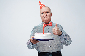 Waist up portrait of satisfied and festively dressed old man in birthday cap showing thumbs-up and holding cake with candle against white background