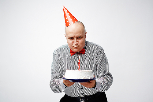 Displeased man in birthday cap looking at burning candle on cake