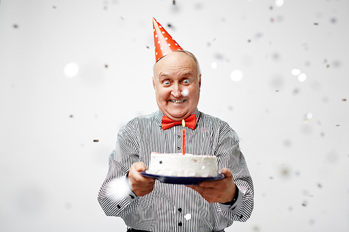 Mature man in birthday cap looking at burning candle in cake
