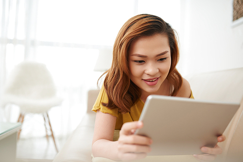 Pretty Asian woman lying on sofa and watching movie on tablet