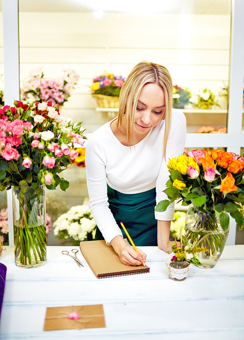 Beautiful young female florist writing something at notebook while standing at her working place