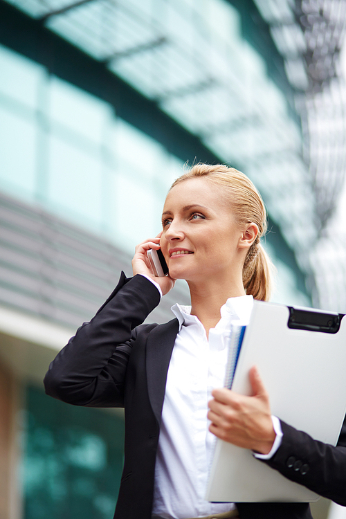 Successful young businesswoman with cellphone calling by modern building