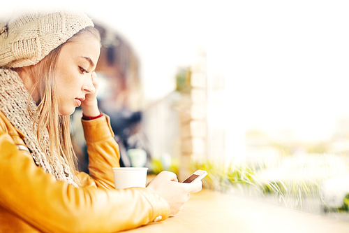 Beautiful sad young girl wearing knitted had, sitting by the window in cafe and looking at her smartphone, waiting for text or call, copy space to the right