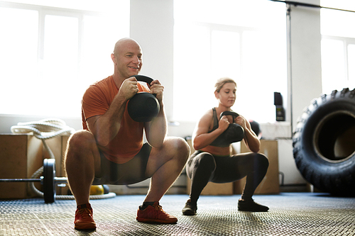 Strong man and woman squatting while exercising with kettle bells