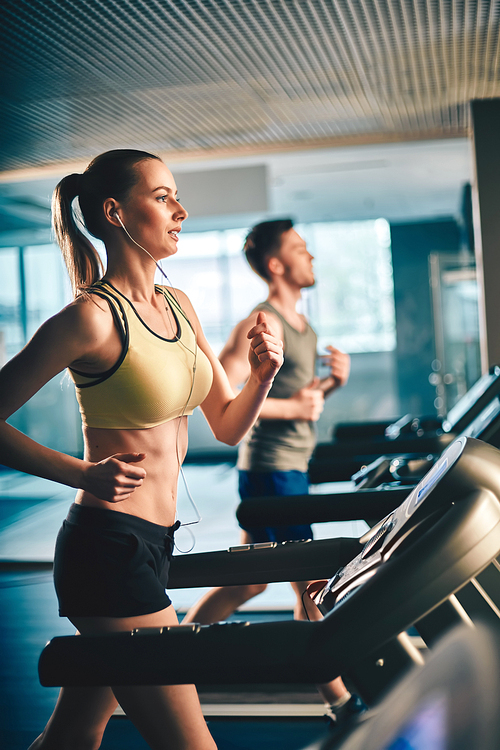 Fit girl with earphones running on treadmill and listening to music with guy on background