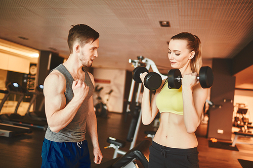 Fit woman with barbells pumping muscles while young trainer consulting her in gym