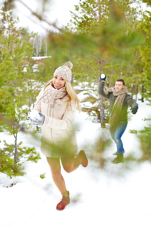 Joyful girl in winterwear playing snowballs with her boyfriend in park or forest