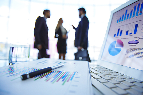 Electronic financial document in laptop and paper with pen above on background of business group talking