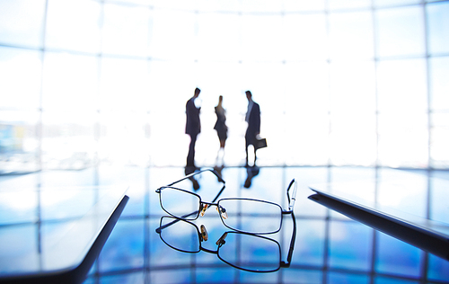 Eyeglasses and touchpads at workplace with businesspeople having meeting on background