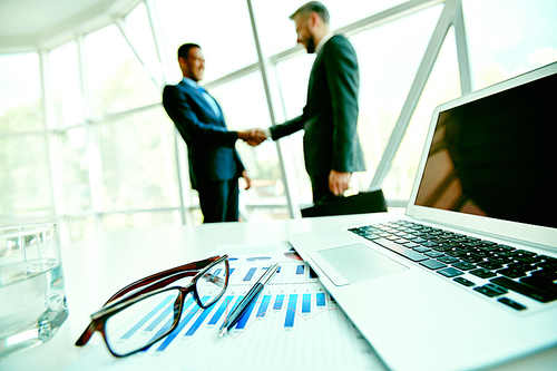 Office stationary items on workplace and handshaking businessmen on background