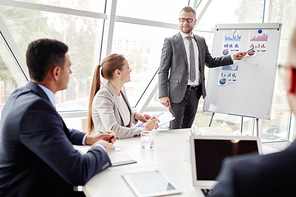 Confident businessman by whiteboard showing data upon sales on the market to his colleagues