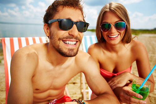 Happy guy in sunglasses and his girlfriend with cocktail resting on beach