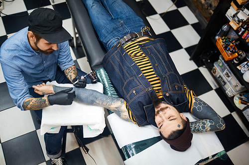 Stylish young man lying in tattoo-salon with master near by