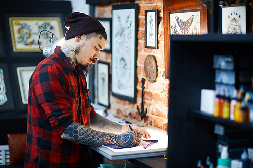 Bearded man in beanie-cap and shirt drawing picture of creative ornament in tattoo-salon