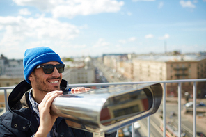 Portrait of young solo tourist smiling while enjoying panoramic view of city from rooftop platform using coin-operated binoculars