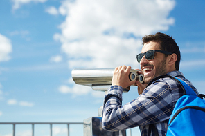 Portrait of young handsome man smiling happily while standing on rooftop against clear blue sky and using coin operated binoculars on viewing platform