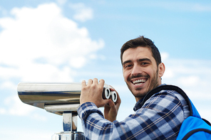 Portrait of young handsome man smiling happily  while using coin operated binoculars  on rooftop viewing platform against clear blue sky