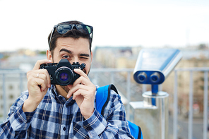 Portrait of handsome young man taking picture with vintage photo camera while standing on rooftop viewing platform against panoramic city view and coin-operated binoculars in background