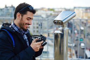 Side view portrait of modern solo tourist  using photo camera to browse pictures while standing on rooftop viewing platform against panoramic city view and coin-operated binoculars in background