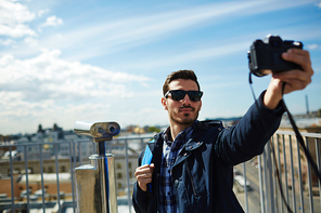 Portrait of handsome young man taking selfie photo while standing on rooftop viewing platform against panoramic city view and coin-operated binoculars in background