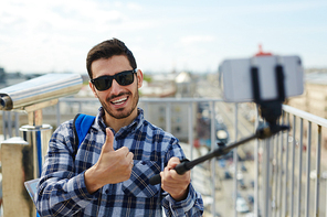 Portrait of handsome young man showing thumbs up to camera taking selfie photo while standing on rooftop against panoramic city view and coin-operated binoculars in background