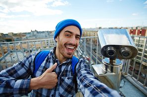Portrait of happy young man showing thumbs up to camera taking selfie photo while standing on rooftop against panoramic city view and coin-operated binoculars in background