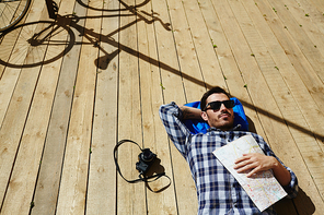 Above view of handsome young man lying on wooden dock planks in sunlight holding map, camera and bicycle nearby