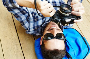 Above view of handsome young man lying on wooden dock planks holding photo camera , enjoying vacation trip