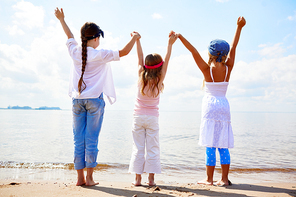 Back view of three ecstatic girls holding by hands while raising them in front of the sea